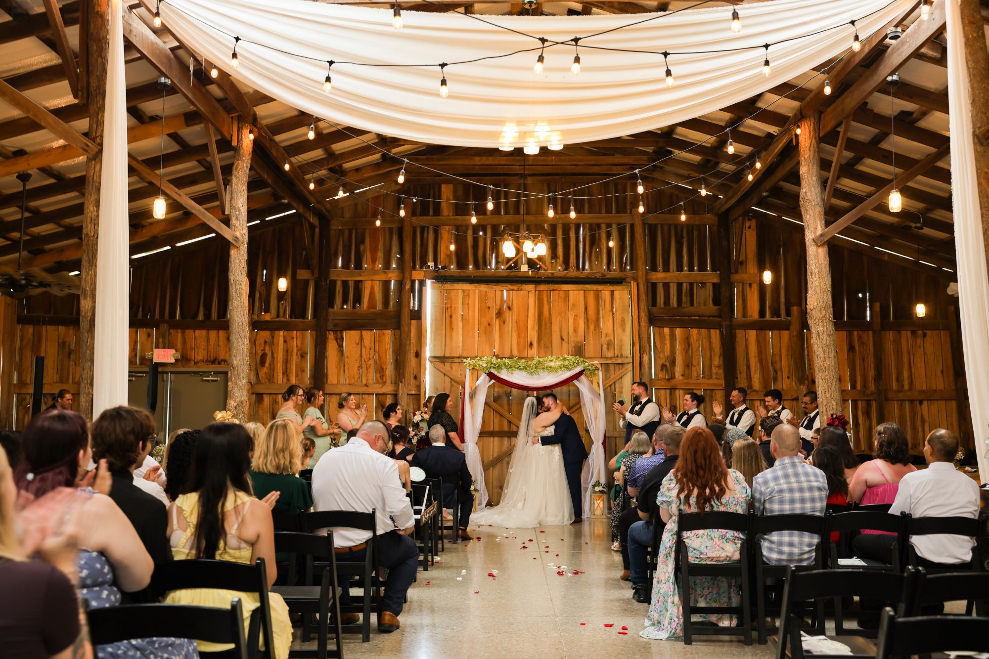 A couple kissing at the end of the aisle under a draped arbor inside the barn