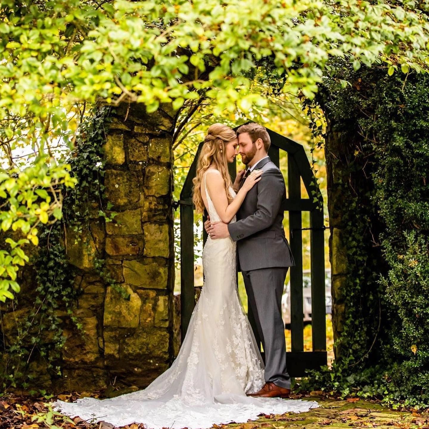 A bride and groom embracing forehead to forehead beside an ivy-covered stone wall and iron gate at Land O' Goshen