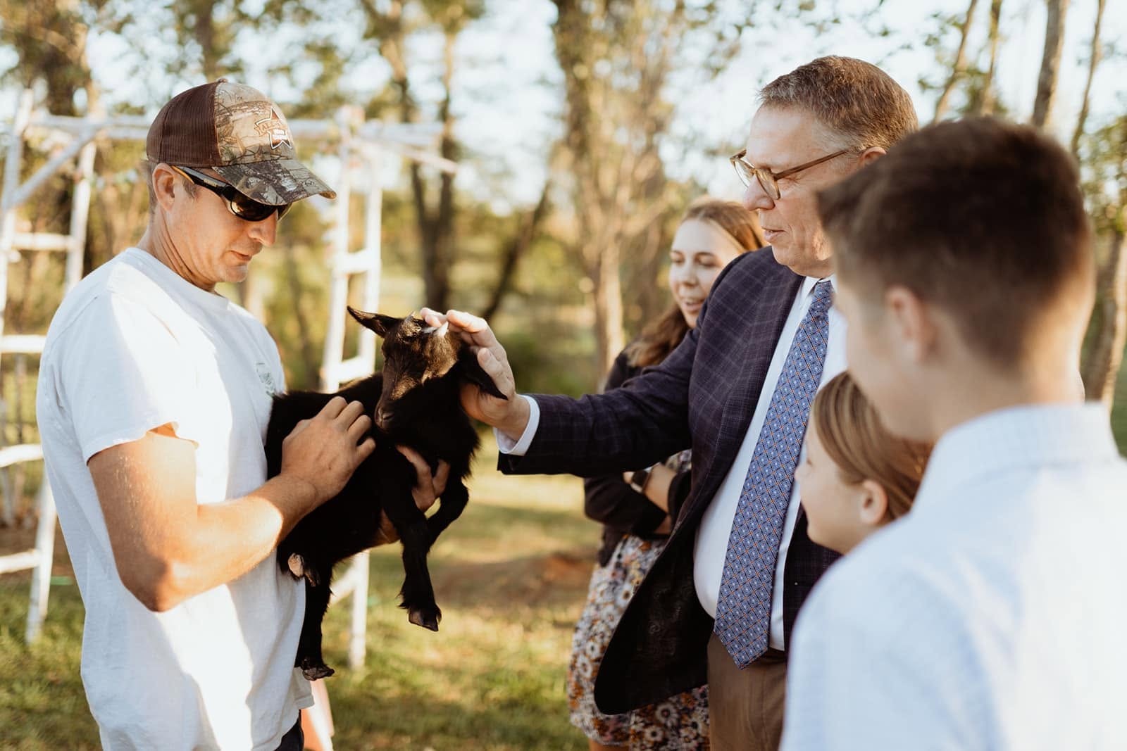 Ian holding a small black goat while wedding guests stop to pet it on the farm